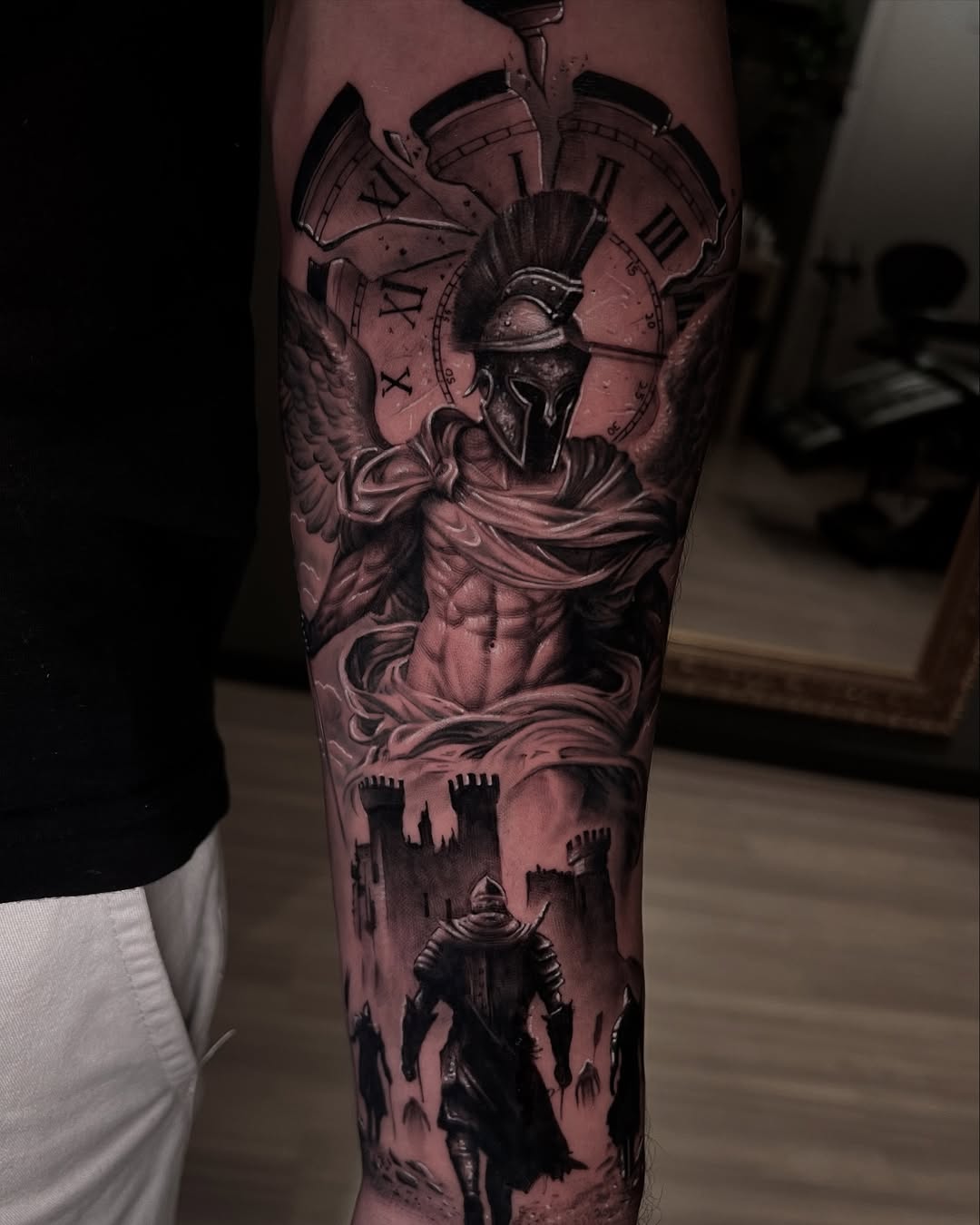 Winged Spartan warrior beneath a Roman clock, tattooed in black and grey realism on a forearm
